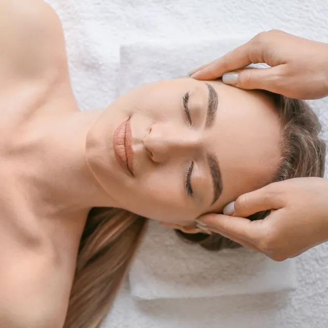A beautician massages the temples of a client lying with her eyes closed, wrapped in white towels