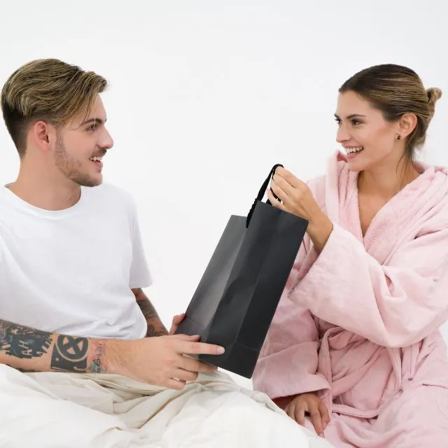 Un homme offre un sac cadeau à une femme en peignoir rose, qui le reçoit en souriant