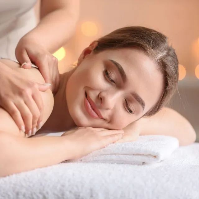 A therapist massages the shoulders of a smiling client lying down, by candlelight