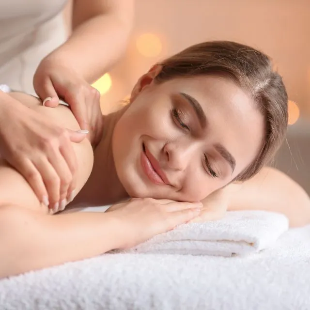 Une praticienne masse les épaules d’une cliente allongée et souriante, à la lueur des bougies